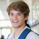 high school senior boy smiles while standing near locker and wearing backpack